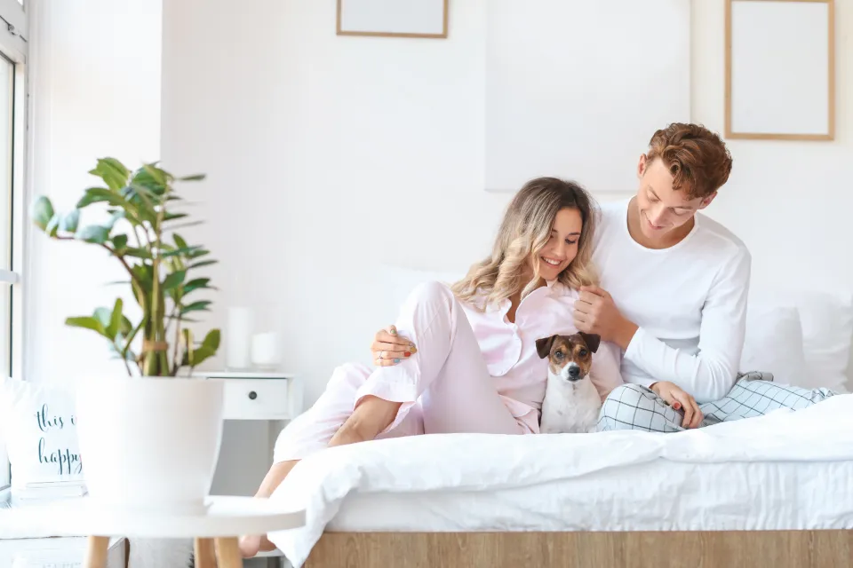 Pareja joven sonriendo en casa, sentados en la cama con su perro